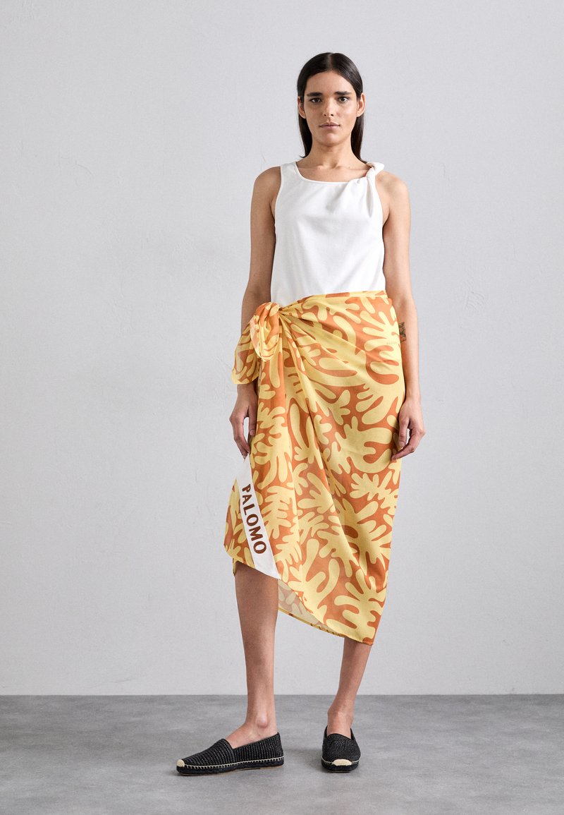 Flowing yellow skirt with orange abstract patterns, tied at the waist, featuring a white label with "PALOMO." Paired with a white top and black shoes.