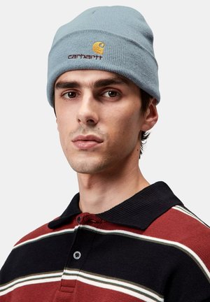Jasnoniebieska dzianinowa czapka z haftowanym pomarańczowym logo. Model nosi koszulkę polo w paski w kolorze czerwonym, czarnym i zielonym, z czarnym, prążkowanym kołnierzem.
