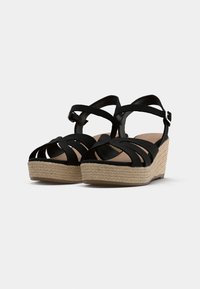 Sandales compensées noires avec brides croisées, détails en jute style espadrille sur la semelle, et finition lisse mate. Fermeture à boucle sur la bride.