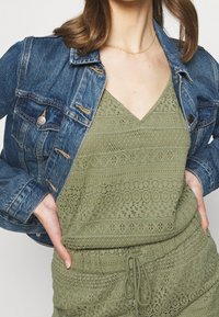 Džínová bunda s měděnými akcenty přes světle olivově zelený, texturovaný, bezrukávový top s výstřihem do V a složitými výšivkami.