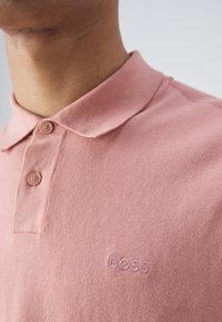 Primo piano di una persona che indossa una polo rosa con colletto, due bottoni e "BOSS" ricamato sul petto.