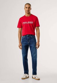 Camiseta vermelha de algodão com texto de logotipo branco, combinada com jeans azuis de ganga e sapatilhas brancas. Design simples, mangas curtas, corte reto.