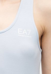 Ανοιχτό μπλε φανελάκι με λευκό λογότυπο "EA7 Emporio Armani" που φοριέται στον ακάλυπτο ώμο και το πάνω μέρος του στήθους.
