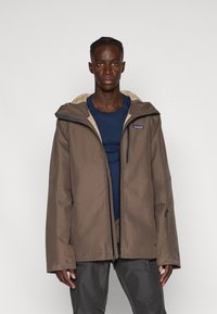 Giacca impermeabile marrone con interno beige, cappuccio e tasche con zip, indossata sopra una maglietta blu e abbinata a pantaloni grigio scuro. Maniche regolabili.