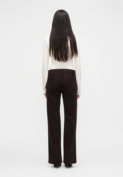 Ženska z dolgimi temnimi lasmi, oblečena v belo rebrasto majico z dolgimi rokavi in temno rjave visokopasne hlače s ravnimi hlačnicami, stoji obrnjena stran.