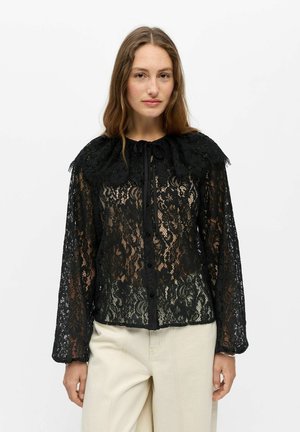 Femme aux longs cheveux bruns portant un chemisier noir en dentelle boutonné avec un grand col et un pantalon crème à jambes larges, debout devant un fond uni.