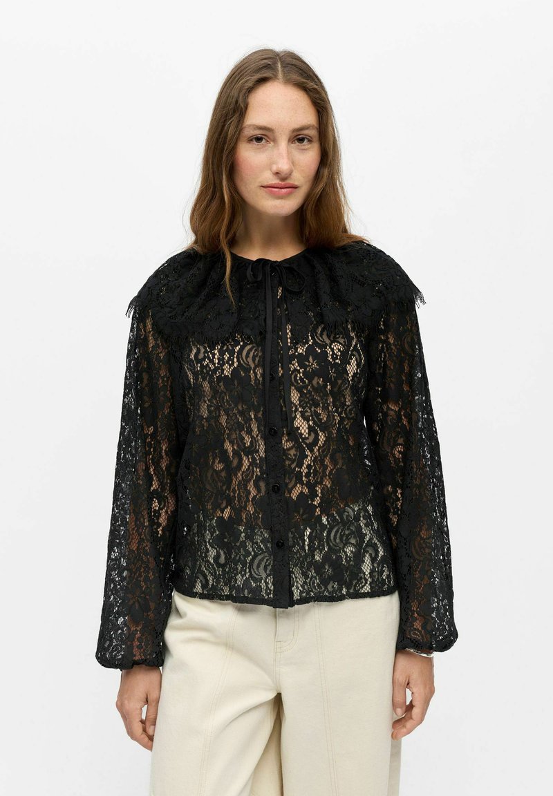 Femme aux longs cheveux bruns portant un chemisier noir en dentelle boutonné avec un grand col et un pantalon crème à jambes larges, debout devant un fond uni.