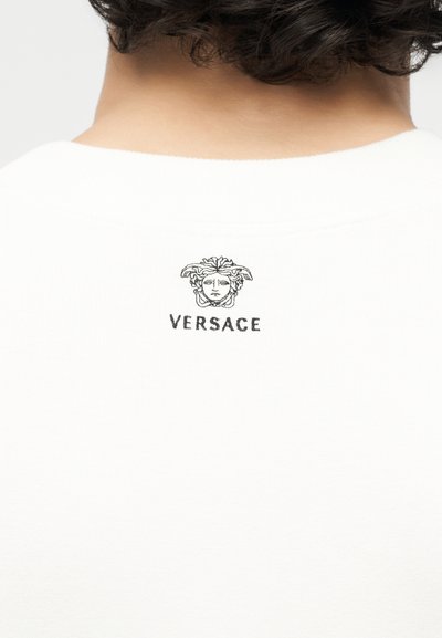 Λαιμός και άνω μέρος της πλάτης ενός ατόμου που φοράει λευκό πουκάμισο με μικρό μαύρο λογότυπο Versace με κεφάλι Μέδουσας στον αυχένα.