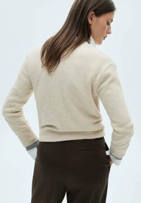 Maglione corto beige realizzato in materiale morbido con orlo a coste. Presenta maniche lunghe e una vestibilità casual. Indossato sopra pantaloni marroni scuri.