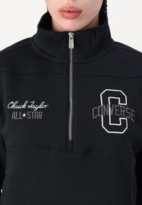 Sudadera con cremallera negra y cuello alto. Presenta bordados en blanco y gris de los logotipos "Chuck Taylor All-Star" y "CONVERSE". Textura suave.