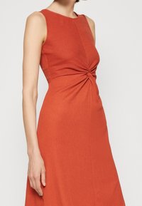 Robe sans manches couleur orange rouille avec un tissu texturé, présentant un détail de torsion à l'avant qui se resserre à la taille, créant une silhouette ajustée.
