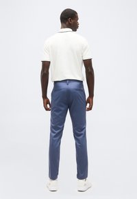 Άνδρας με πλάτη, φορώντας λευκό πόλο μ ε κοντά μανίκια, μπλε παντελόνι και λευκά αθλητικά παπούτσια, σε απλό φωτεινό φόντο.