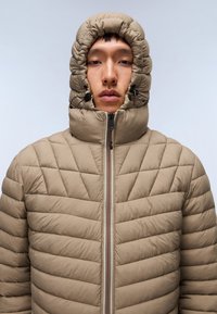 Giacca a vento in khaki con colletto alto e cappuccio con cordino. Presenta una texture trapuntata e una cerniera a contrasto.