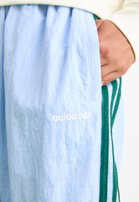 Gaiši zila adidas sporta bikses ar balto logo, ar zaļām sānu joslām un strukturētu, vieglu audumu. Sānos redzama kabata.