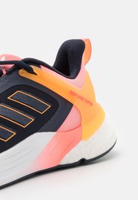 La chaussure de sport noire et rose présente une tige texturée, des accents orange, et une semelle blanche rembourrée ; elle inclut un logo "RESPONSE SUPER".