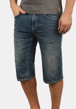Homme portant un short en denim bleu délavé avec les mains dans les poches, associé à un t-shirt gris, montré de la taille aux genoux.