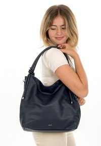 Mladá žena drží na rameni velkou tmavě modrou koženou kabelku, má na sobě bílý krátký rukáv a béžové kalhoty.