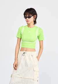Šviesiai žalia, trumpomis rankovėmis, apkarpyta sportinė palaidinė su tinkline detalėmis ir Adidas logotipu. Derinama su šviesiai smėline cargo sijonu.