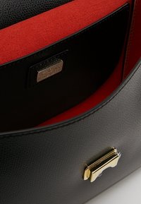Czarna skórzana torba o teksturowanej powierzchni, czerwona zamszowa podszewka, złote akcesoria i dyskretna tabliczka z logo wewnątrz. Gładki, prostokątny kształt.