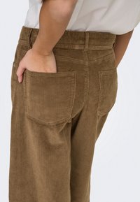 Personne portant un pantalon en velours côtelé marron avec la main droite dans la poche arrière droite sur un fond uni.