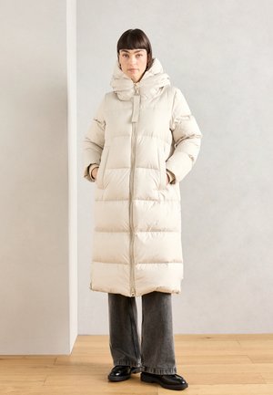 Beige gewatteerde jas met een hoge kraag en ritssluiting, voorzien van een gestikte structuur, twee zijzakken en een relaxte pasvorm. Gecombineerd met grijze jeans.