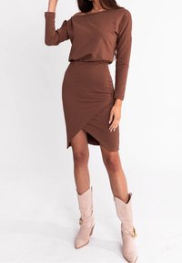 Femme portant une robe portefeuille marron à manches longues associée à des bottes de cowboy beige mi-mollet, debout devant un fond blanc.