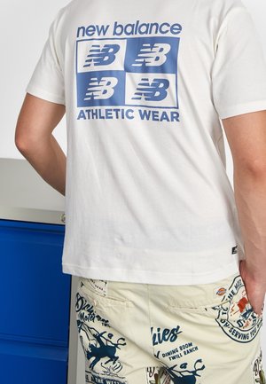 Weißes New Balance T-Shirt mit blauem Logo und dem Text „new balance ATHLETIC WEAR“, kombiniert mit gemusterten, hellen Shorts mit Text und Grafiken.