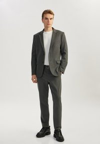 Blazer gris y pantalones a juego de una tela suave, camisa blanca y zapatos negros; diseño estructurado con un único bolsillo en el blazer.