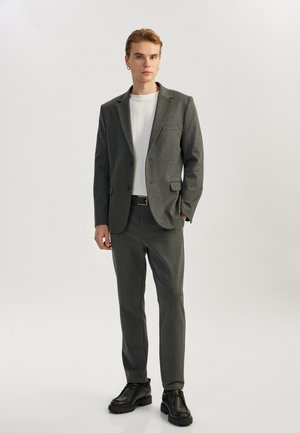 Blazer gris y pantalones a juego de una tela suave, camisa blanca y zapatos negros; diseño estructurado con un único bolsillo en el blazer.