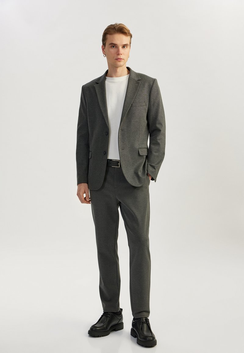 Blazer gris y pantalones a juego de una tela suave, camisa blanca y zapatos negros; diseño estructurado con un único bolsillo en el blazer.