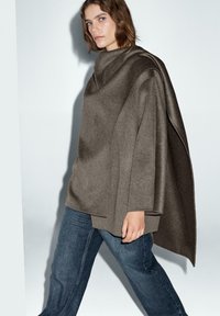 Hnědý pončo z vlněné směsi s volným střihem, sešikmými okraji a jemnou texturou, v kombinaci s tmavě modrými džíny.