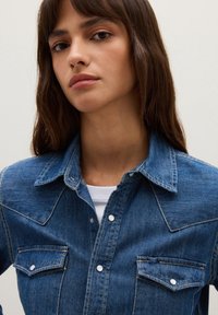 Camicia in denim blu scuro, con colletto a punta, tasche frontali, bottoni a scatto e dettagli di cucitura a contrasto. Chiusura con bottoni al colletto.