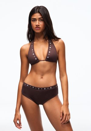 Nuori nainen, jolla on pitkät tummat hiukset, päällään ruskea niitillä koristeltu halter-bikinitoppi ja samaan sarjaan kuuluvat korkeavyötäröiset alaosat, seisoo valkoista taustaa vasten.