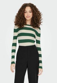 Groene en crèmekleurige gestreepte ribbeltrui met lange mouwen, gecombineerd met hoge zwarte broeken. De trui heeft een aansluitend ontwerp met contrasterende kleuren.