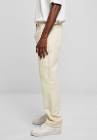 Jeans de couleur claire, à jambe droite, en coton avec une surface texturée. Comprend des poches et un matériel en métal basique. Portés avec des baskets blanches.
