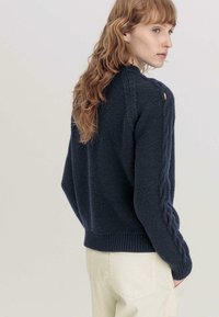 Marineblauer Strickpullover mit strukturiertem Stoff, Kabelmuster entlang der Ärmel und geripptem Saum; getragen mit hellen Hosen.