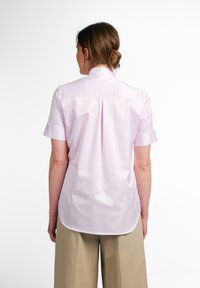 Camicia a maniche corte in rosa chiaro, realizzata in cotone morbido, con colletto a punta e orlo leggermente curvo. Dettaglio della piega sul retro visibile.