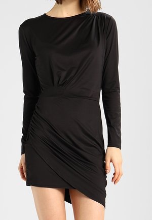 Woman wearing a black long-sleeve dress with an asymmetrical ruched skirt and a fitted waist, standing against a plain background.