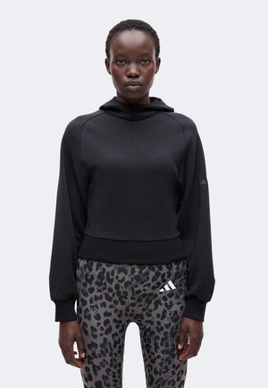 Lühikeste juustega noor inimene, kes kannab musta kapuutsiga pusa ja hallide leopardimustriliste retuusidega, millel on valge Adidas logo, seisab ühetaustal.