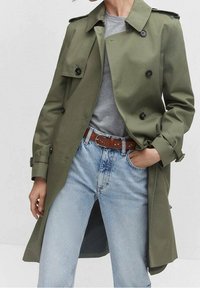 Olivgrüner Trenchcoat mit doppelseitiger Knopfleiste, Schulterklappen und weicher Textur, kombiniert mit hellblauen Jeans und einem grauen Hemd.