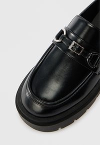 Black leather loafer with a shiny finish. Features a silver buckle and strap detail. Thick black sole with a textured edge for grip.