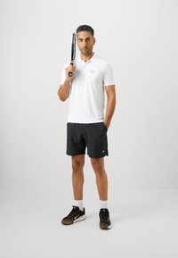 Homem em polo branco e calções pretos segura uma raquete de ténis sobre o ombro, de pé com uma mão no bolso contra um fundo branco liso.