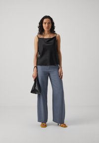 Black satin camisole with a cowl neckline, paired with light blue wide-leg trousers. Holds a black bag and wears yellow block-heel sandals.