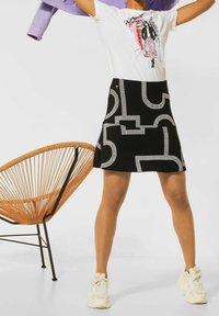 Kvinna som bär en svart mönstrad kjol, en vit grafisk t-shirt, vita sneakers, hållande en lila jacka över huvudet, stående nära en brun vävd stol.