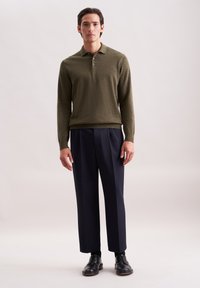 Uomo in piedi che indossa un maglione polo a maniche lunghe verde oliva, pantaloni larghi blu navy, calze nere e scarpe eleganti in pelle nera su uno sfondo chiaro.