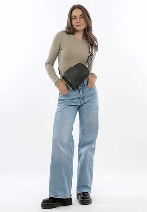 Sac en cuir noir à porter en bandoulière, avec un dessus arrondi, une surface texturée et une bandoulière ajustable, porté sur un haut à manches longues taupe et un jean bleu à jambes larges.