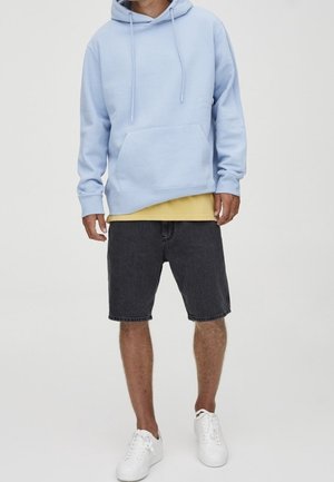 Mann trägt hellblauen Kapuzenpullover, gelbes Shirt darunter, schwarze Jeans-Shorts und weiße Turnschuhe, steht vor einem schlichten hellen Hintergrund.