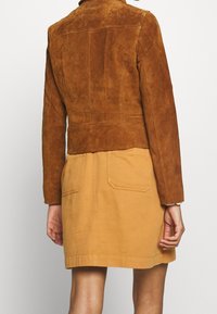 Braune Wildlederjacke mit gekürztem Saum und Kragen, kombiniert mit einem hellbraunen Rock mit zwei Gesäßtaschen und einer glatten Textur.