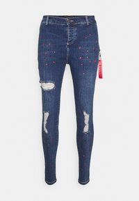 Jeans ajustados de color azul oscuro con agujeros en las rodillas, manchas de pintura rojas y negras, y una etiqueta rosa de SikSilk adjunta al bolsillo derecho.