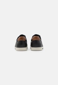 Zapatillas negras con diseño texturizado, acentos de cuero marrón en el talón y suelas de goma blanca. Vista desde atrás, que muestra las características bicolor.